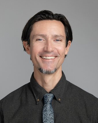 A man with dark hair and a goatee is smiling at the camera. He is wearing a dark button-up shirt and a patterned tie, with a neutral gray background.