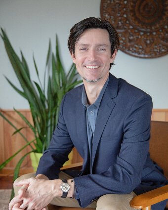 A man with dark hair and a goatee is sitting in a wooden chair, smiling confidently. He is wearing a navy blazer over a grey shirt, with a plant in the background.