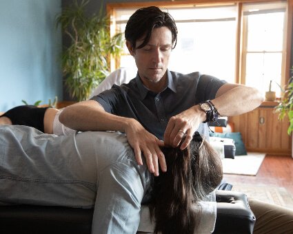 A man with dark hair is performing a chiropractic adjustment on a person lying face down on a treatment table. The setting includes plants and natural light from windows in the background.