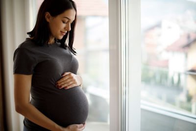 A pregnant woman gently holds her belly while standing by a window.